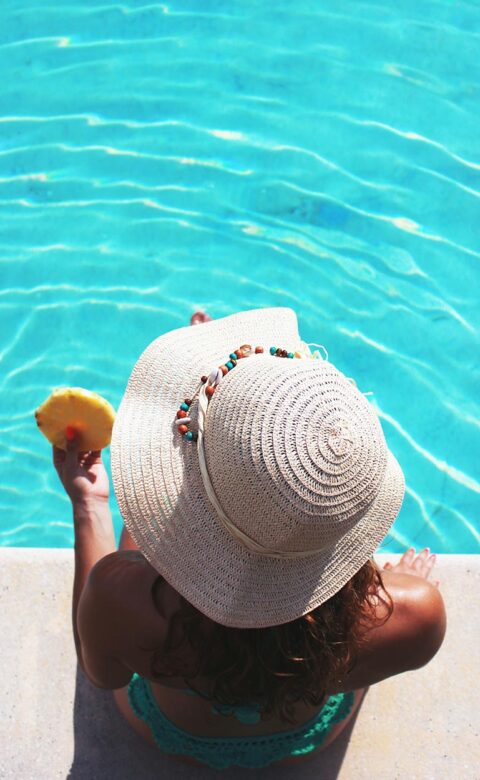 woman in large sun hat holding pineapple slice sitting with legs in pool