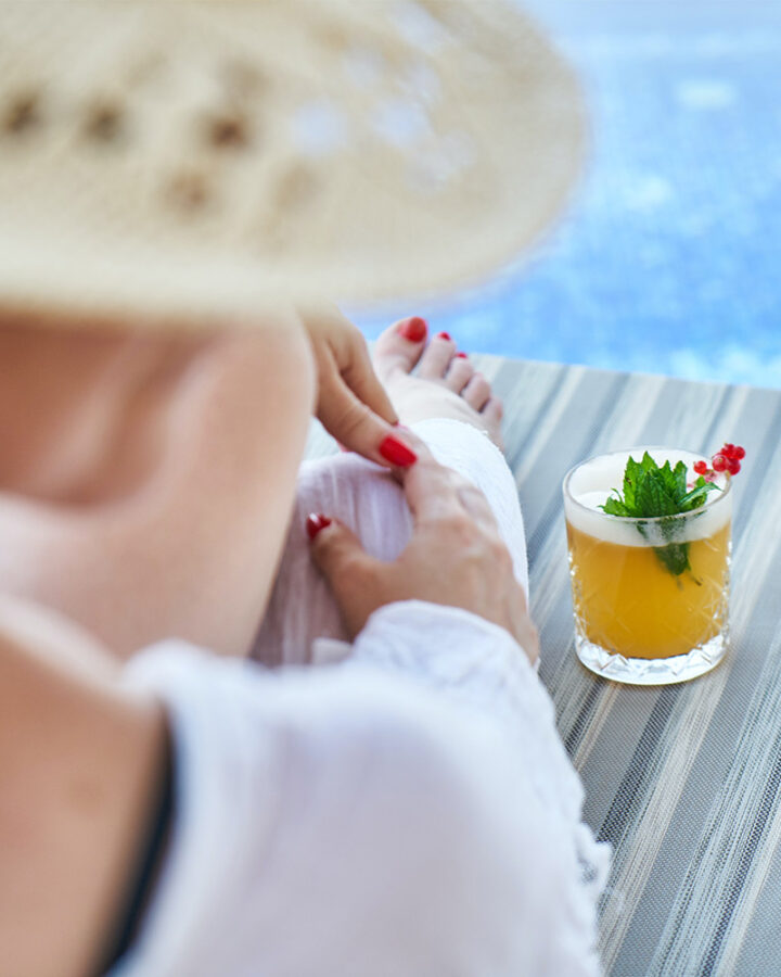 woman sits on chaise lounge next to pool and cocktail